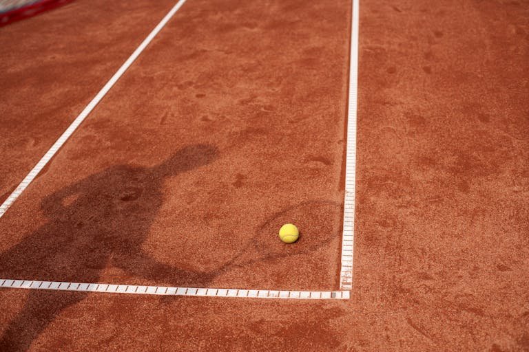 テニスコート（クレーコート）上で、黄色いテニスボールとともに映る選手の影。スポーツやテニスをテーマにした用途に最適。